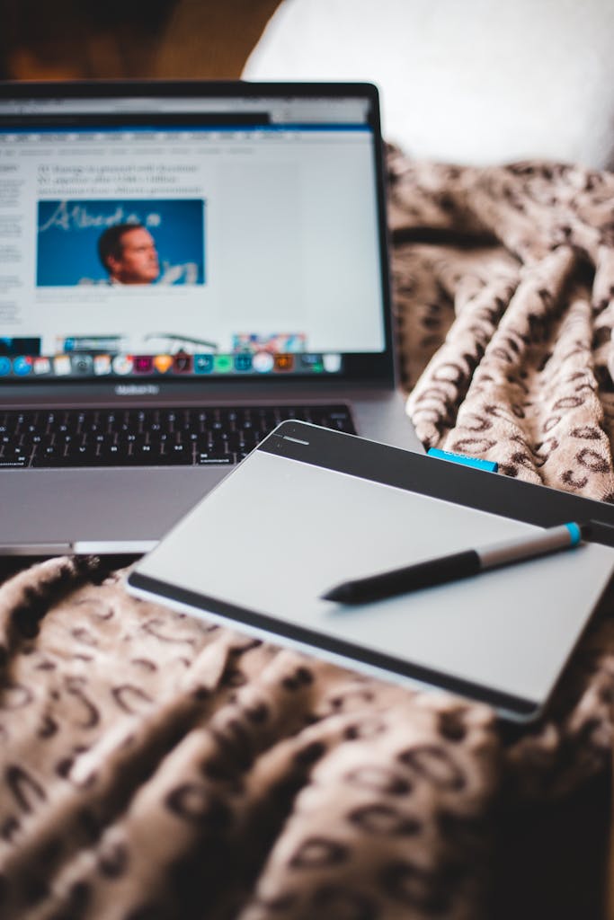 From above of open netbook with picture on screen near notebook with empty sheet of paper with pen on crumpled plaid on bed with pillow at home